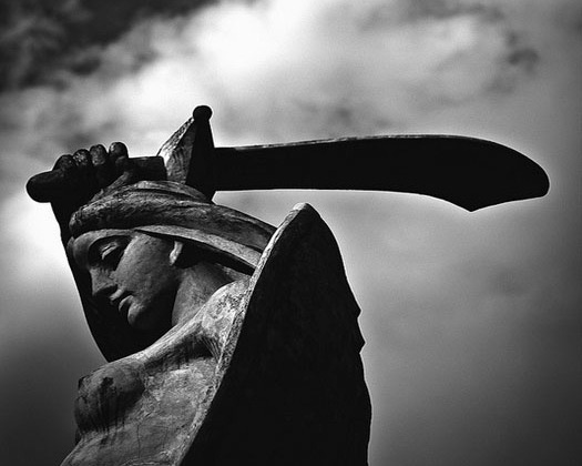 A weathered stone statue holding a sword against a cloudy sky